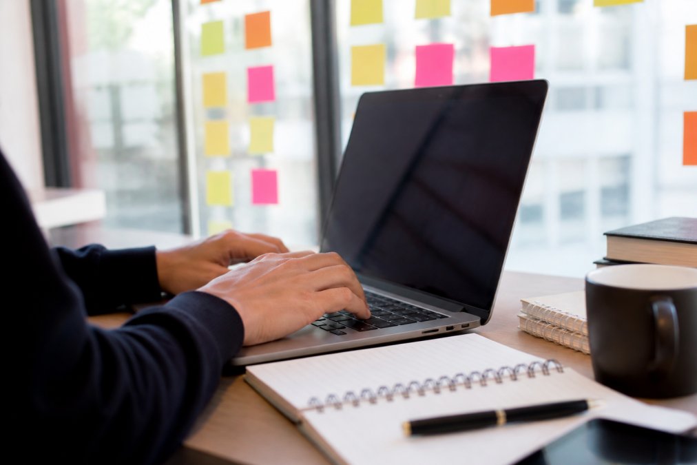 Close-up of hands typing on a laptop with a mind-mapping application visible on screen, sticky notes arranged on a glass wall in the background