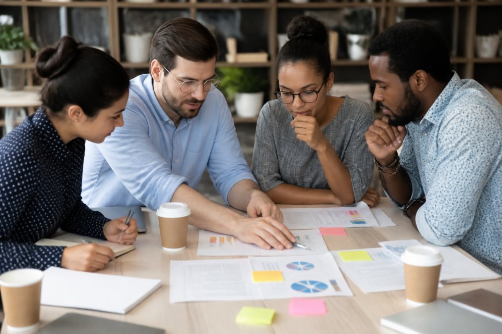 Business team reviewing customer data and market research on multiple screens in a collaborative workspace, with charts and metrics visible on monitors, natural office lighting