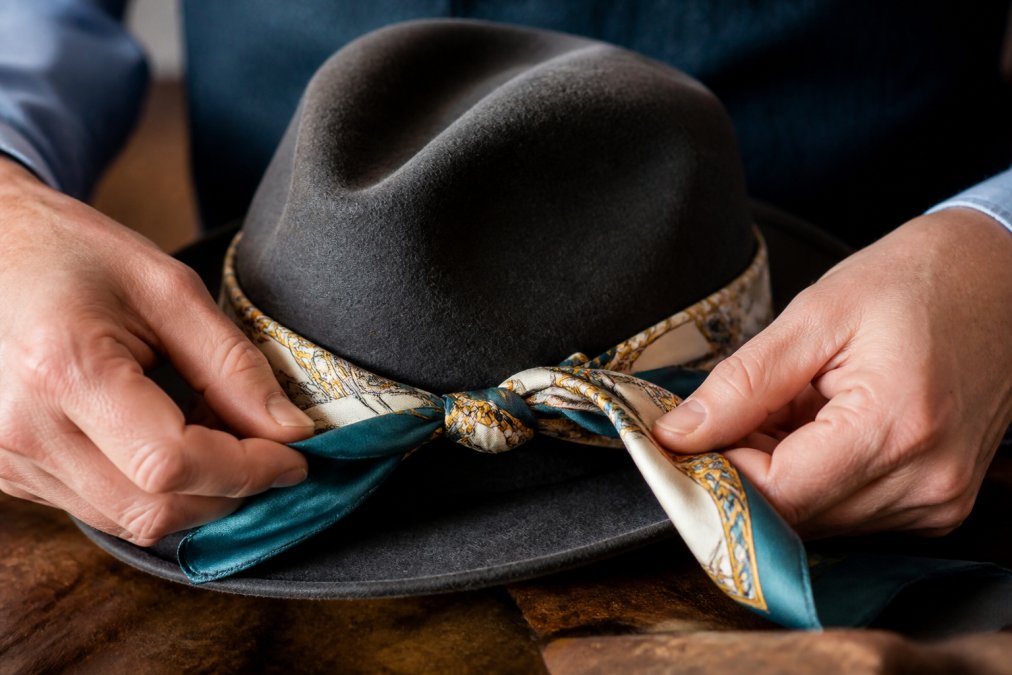 Close-up of hands tying a silk scarf around the brim of a felt fedora hat, showing the knotting technique with soft natural lighting