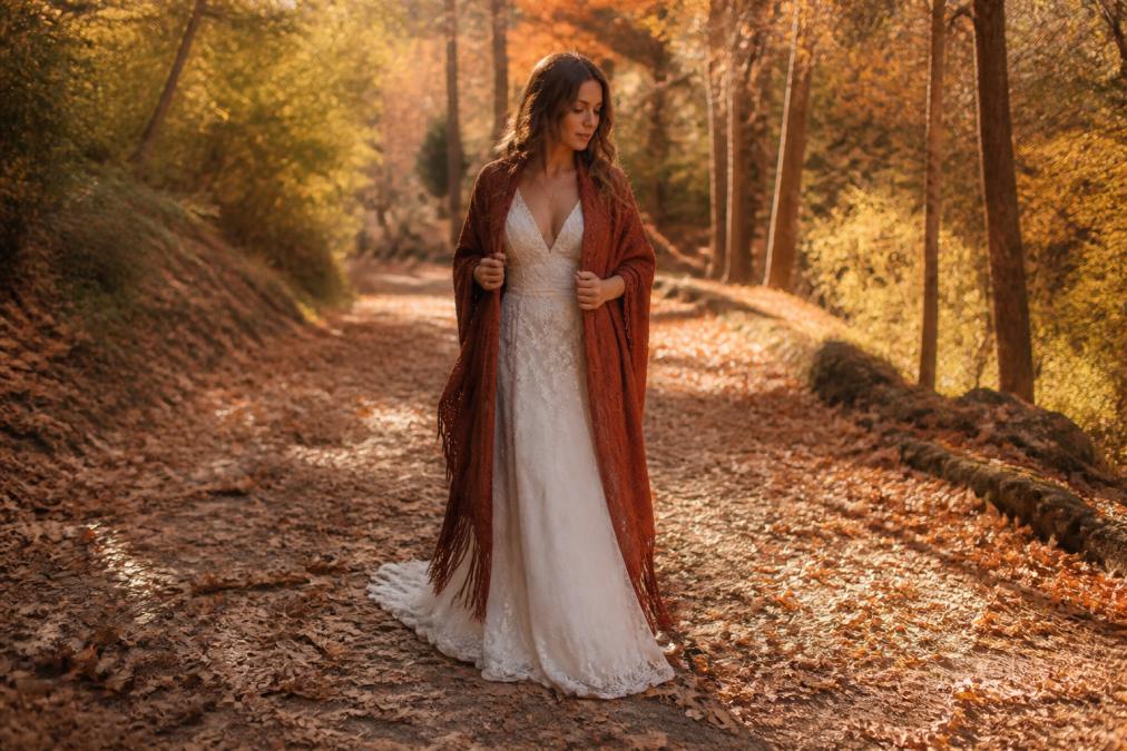 A bride wearing a flowing bohemian wedding gown with a fringed rust-colored ruana draped over her shoulders, standing on a leaf-covered path in an autumn outdoor setting with soft golden afternoon light