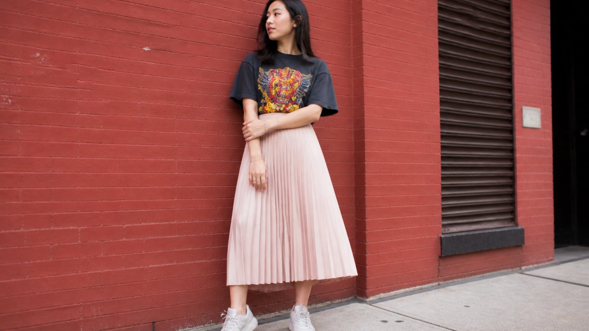 A young adult wearing a bold graphic tee tucked into a pleated midi skirt with sneakers, standing against a brick wall in an urban setting, natural daylight, candid street style shot