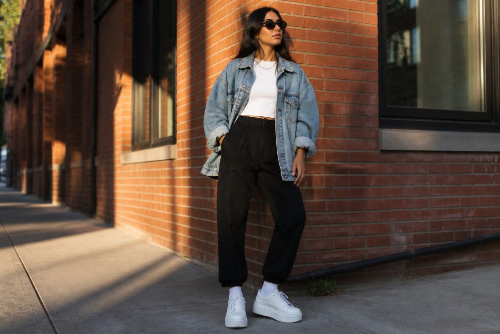 A woman styled in layered streetwear with an oversized vintage denim jacket, fitted white base layer, black tapered pants, and bold white platform sneakers, photographed in an urban setting with natural afternoon sunlight casting shadows on brick buildings