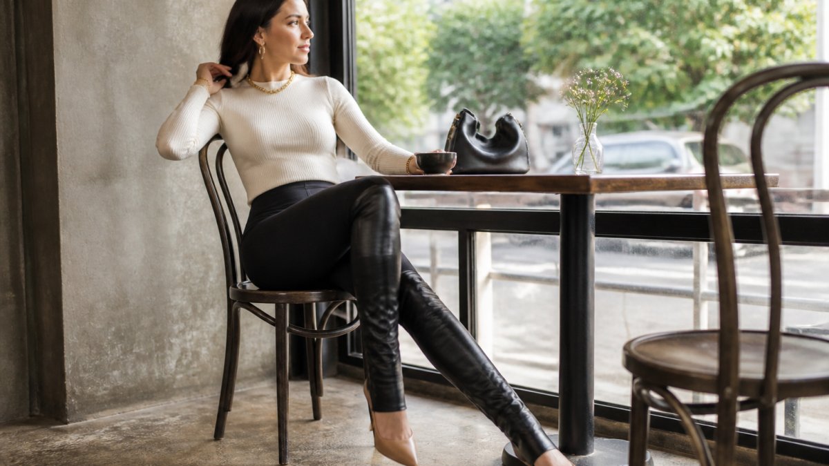 Woman wearing black leggings with thigh-high boot sleeves over nude heels, styled with a cream-colored fitted sweater and gold jewelry, seated in a modern café setting with natural window lighting