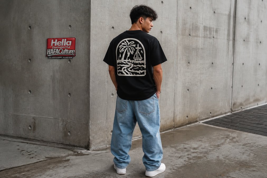 Young person in an island-inspired graphic tee and baggy denim posing against an urban concrete wall.