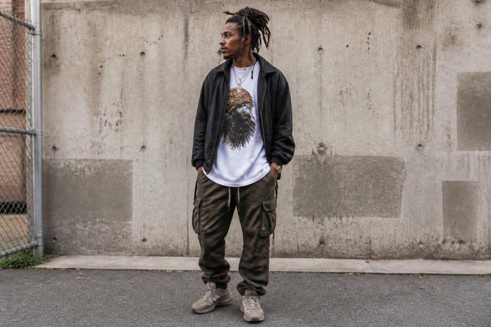 Person wearing layered conscious streetwear with an oversized graphic tee under an open unstructured jacket, demonstrating texture contrast and silhouette proportion in an urban outdoor setting with natural daylight