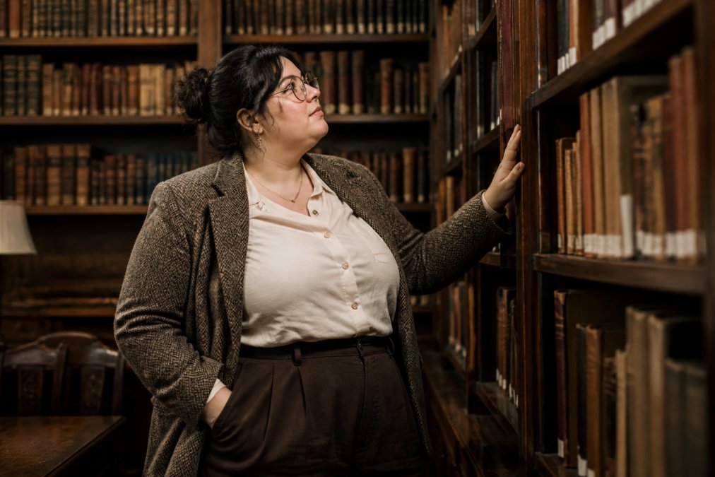 A plus-size woman with a curvy figure wearing a tweed blazer, button-front blouse, and high-waisted trousers, standing in a cozy library with bookshelves
