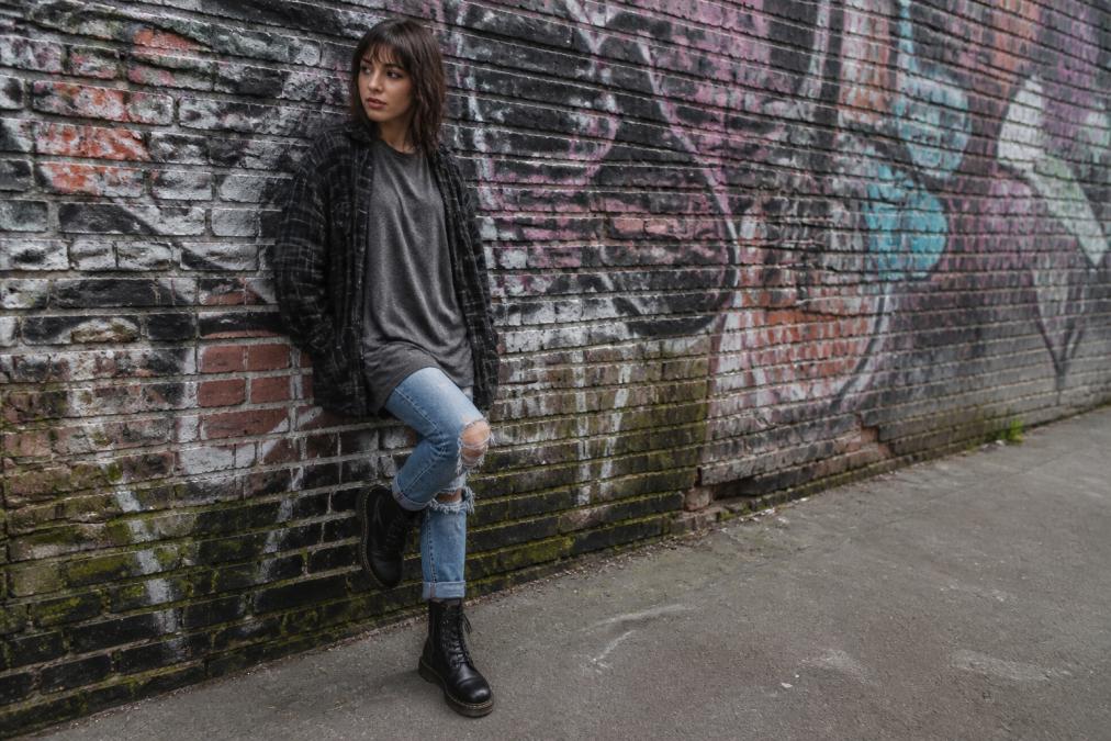 A person wearing an oversized graphic tee under an open black flannel shirt with ripped jeans and combat boots, standing against a graffiti-covered brick wall in an urban setting during overcast daylight