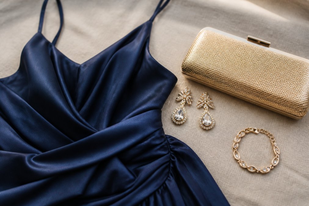 Close-up of formal dress styled with elegant statement earrings, delicate bracelet, and small clutch arranged on a neutral surface, showing complete accessory coordination in soft natural light