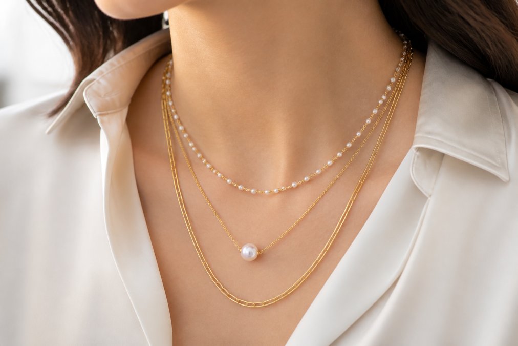 Close-up of a woman's neckline showing multiple layered pearl and gold chain necklaces at varying lengths, worn with a silk blouse, demonstrating modern effortless elegance