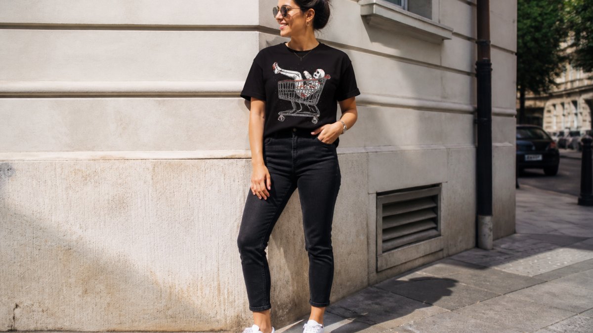 Person wearing a bold graphic tee tucked into fitted dark jeans with white trainers, standing casually outdoors with natural daylight illuminating the outfit