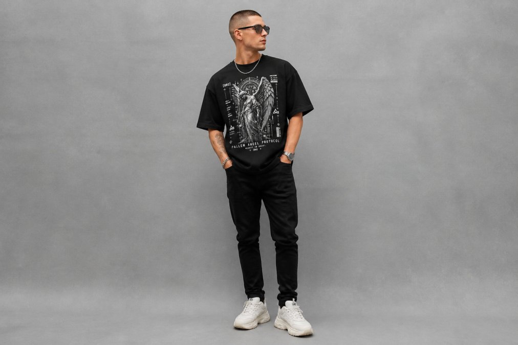 Person wearing an oversized heavyweight graphic tee in dark color with tapered black denim and white chunky sneakers, standing against a neutral gray backdrop showing balanced proportions between loose top and fitted bottom