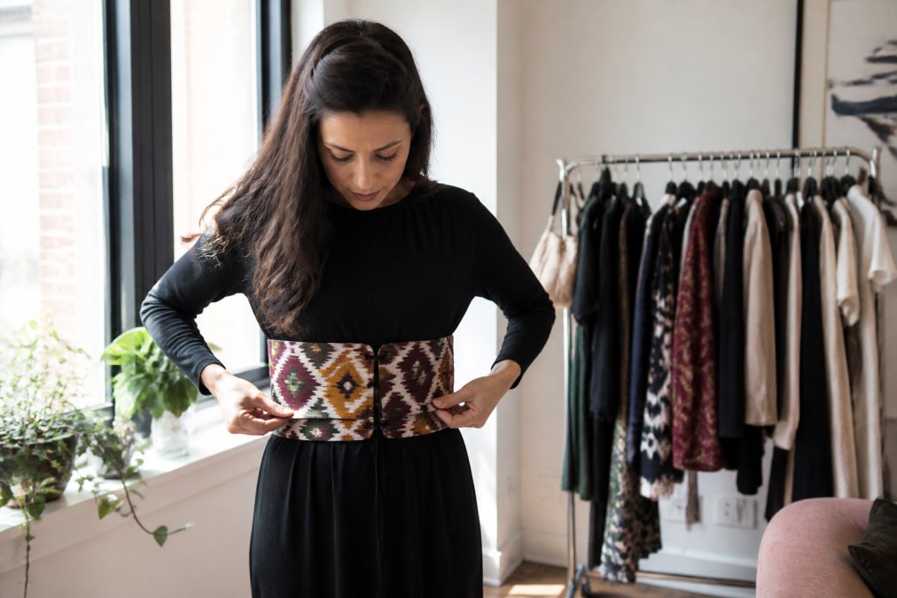 Woman in a bright apartment adjusting an ikat corset belt over a solid black dress, morning light through tall windows, clothing rack visible behind her