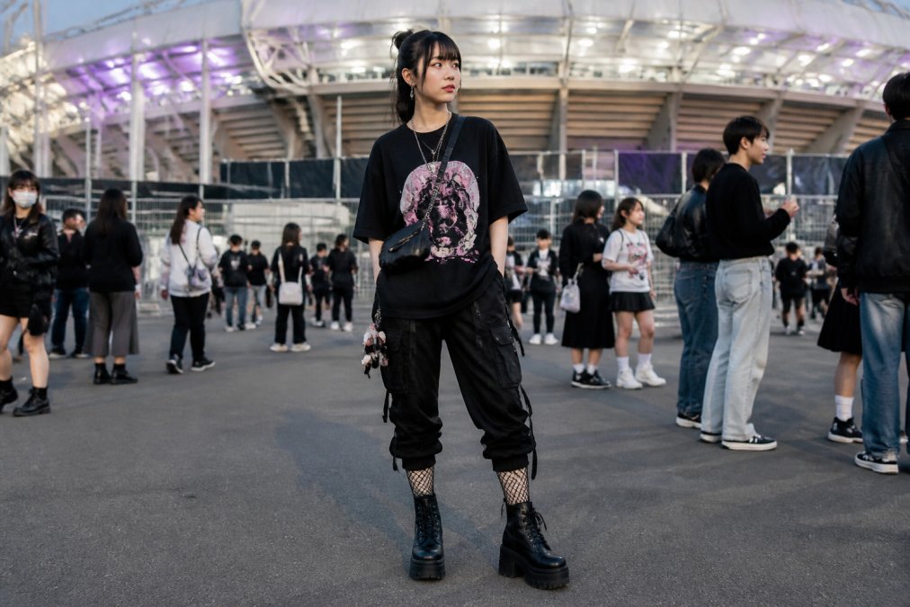 Person wearing an edgy streetwear K-pop concert outfit with oversized black graphic tee, fitted cargo pants with multiple pockets, platform boots, fishnet socks, and a small crossbody bag, standing at an outdoor venue entrance with stadium lights visible in the background
