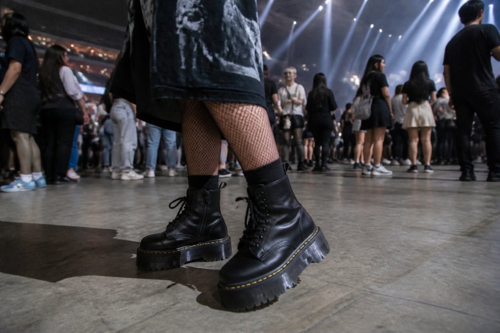 Close-up of feet wearing black platform boots with thick treads, paired with fishnet socks and the hem of an oversized graphic tee, standing on the concrete floor of a concert venue with stage lights visible overhead