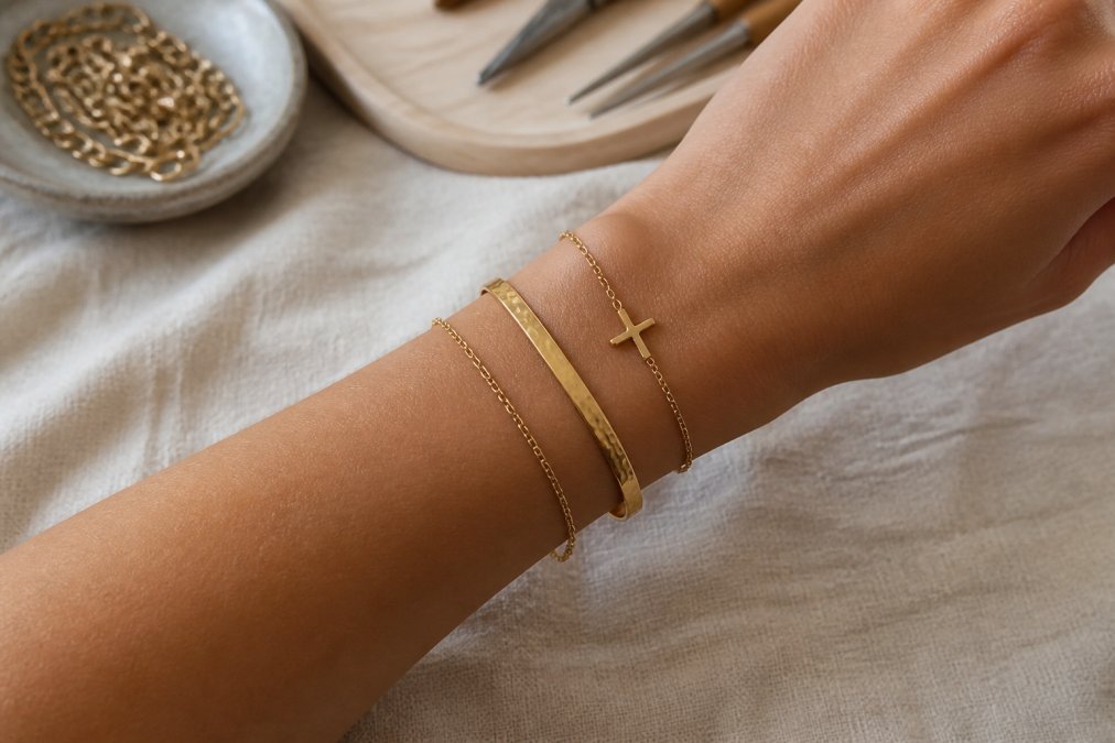 Close-up of a female wrist wearing a layered stack of three gold bracelets: a thin chain, a textured bangle, and a delicate cross bracelet, styled against a neutral linen background in soft natural light