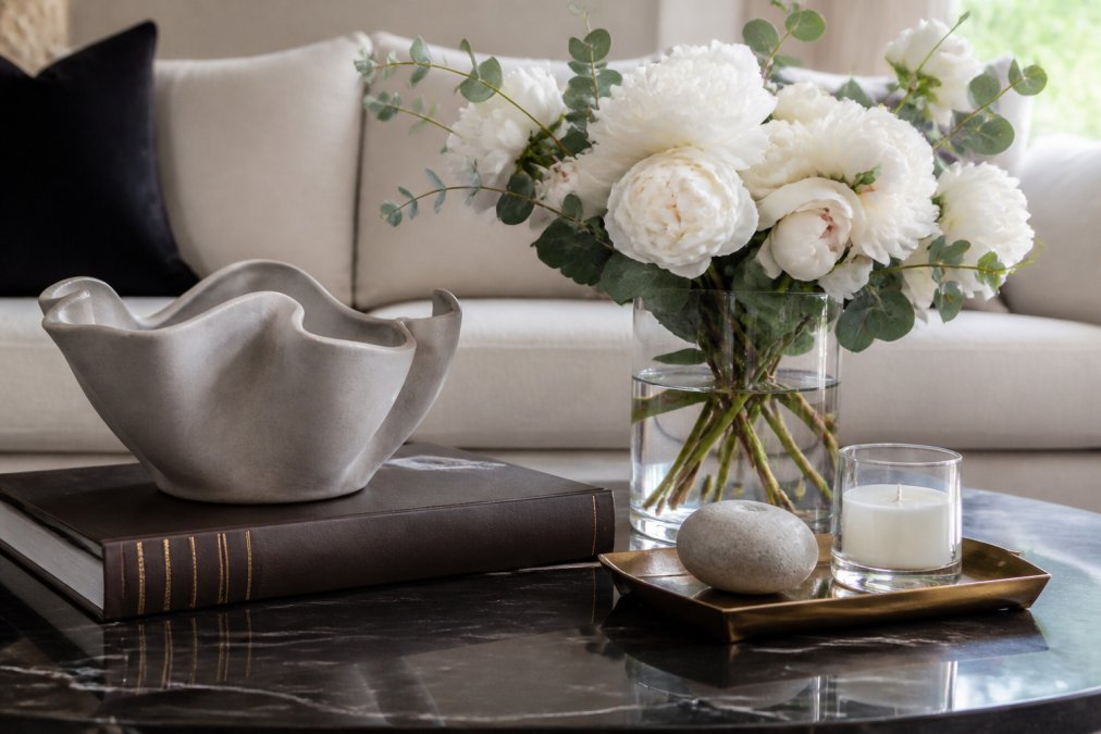Elegantly styled coffee table surface featuring a leather-bound coffee table book with gold-embossed spine, a sculptural ceramic vessel in soft gray with organic, undulating form, a clear glass vase holding fresh white peonies and eucalyptus greenery, and a small brass decorative tray holding a single smooth stone and a luxury candle, arranged with intentional spacing and varying heights