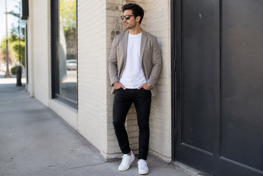 Person wearing a minimalist graphic tee layered under an open neutral-toned blazer with fitted black denim and white sneakers, standing in natural daylight