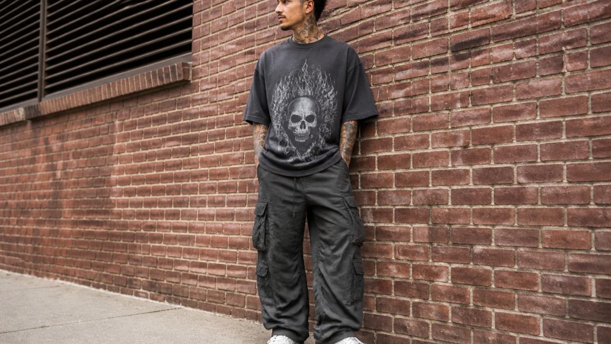 Person wearing an oversized heavyweight graphic tee with baggy cargo pants and chunky white sneakers, standing against an urban brick wall in natural daylight