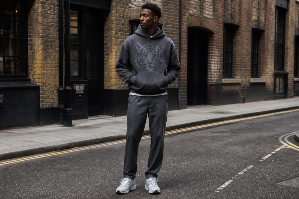 Young man wearing an oversized hoodie styled with tapered trousers and trainers, standing in an urban street setting with natural daylight, relaxed confident posture