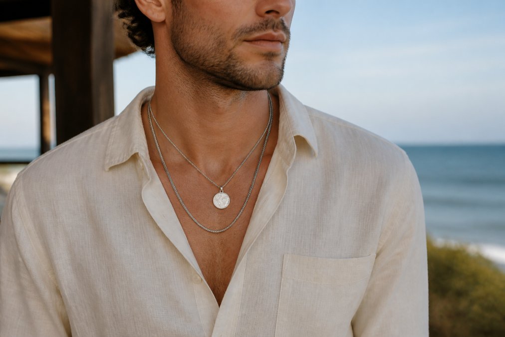 Man wearing two layered necklaces of different lengths, one shorter pendant-style piece with engraving and one longer minimalist chain, styled with a casual cream button-up shirt, showing balanced understated layering in natural daylight