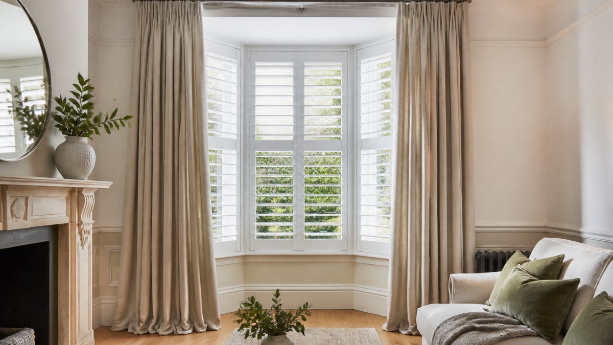 A bay window in a period London home with white plantation shutters and a pair of floor-length linen curtains hanging from a slim metal pole, showing the layering detail