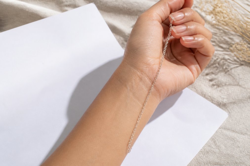 Close-up of a woman's wrist and hand in natural daylight, holding a delicate sterling silver chain against her skin beside a sheet of white paper
