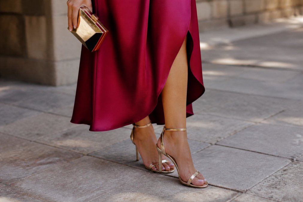 Close-up of a woman's feet and lower legs showing an elegant pair of strappy heels paired with a luxurious jewel-tone statement dress, with a structured metallic clutch visible, demonstrating complete wedding guest styling