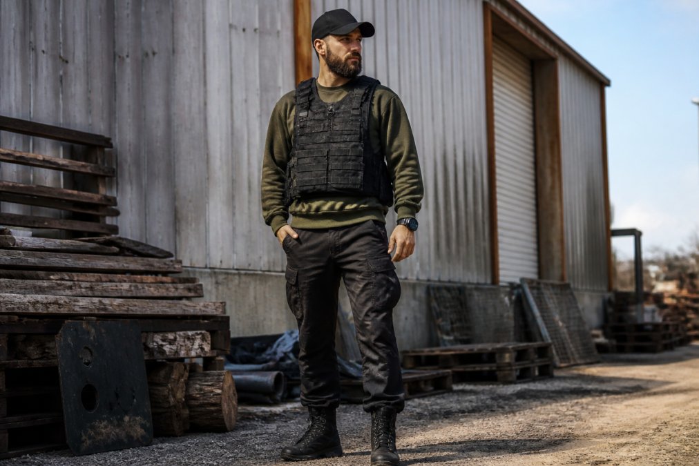 Male model wearing a tactical streetwear outfit with visible layering: heavyweight crewneck under an open tactical vest with MOLLE webbing, cargo pants, and boots, photographed outdoors in natural daylight