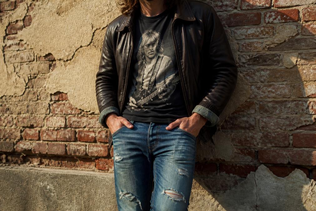 Close-up of a vintage-style band tee layered under an open leather bomber jacket, cuffed sleeves visible, paired with distressed jeans, shot against a weathered brick wall with natural side lighting