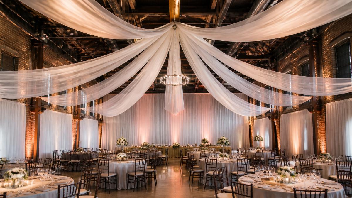 A wedding reception in a converted warehouse with soaring ceilings, white chiffon draping running from a central point outward, soft uplighting on the fabric, round tables with neutral linens below