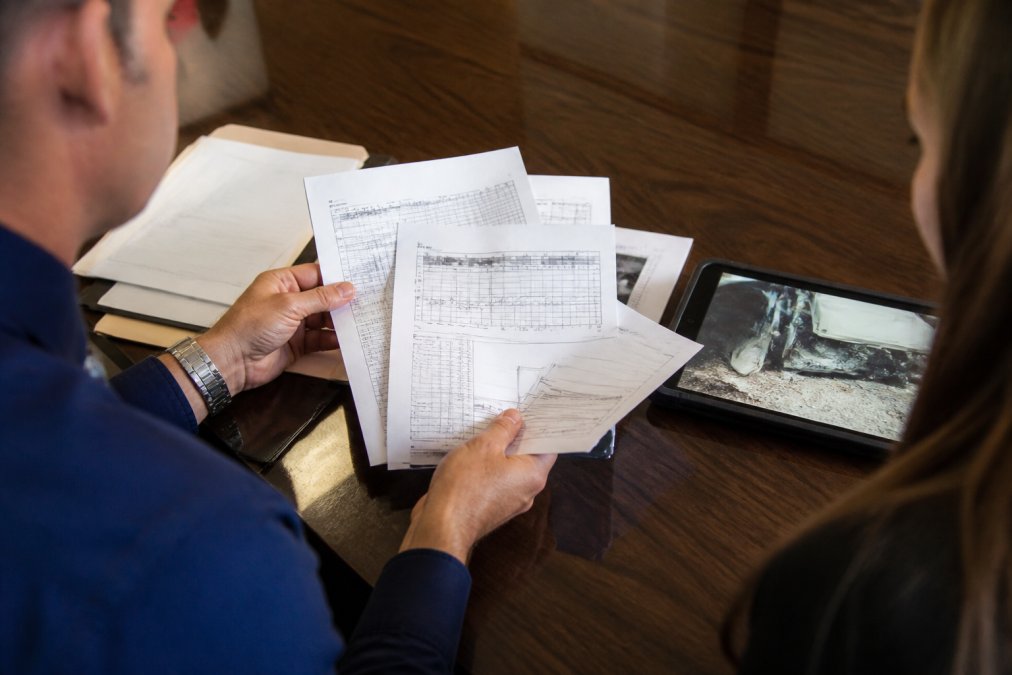 Close-up of hands holding printed electronic logging device records and driver logs on a desk next to tablet displaying accident scene photographs