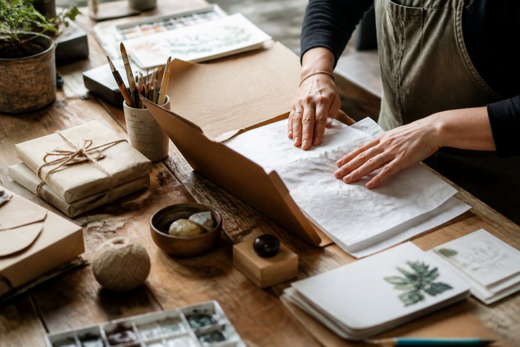 Hands carefully packing a handmade order in a small creative studio, showing personal attention to detail with art supplies and wrapped pieces on a wooden work table