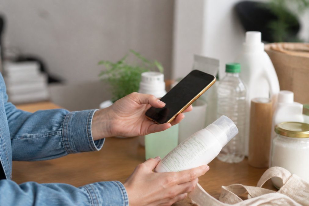 Close-up of hands holding a product package while reviewing sustainability information and certifications on a smartphone screen, cross-referencing claims