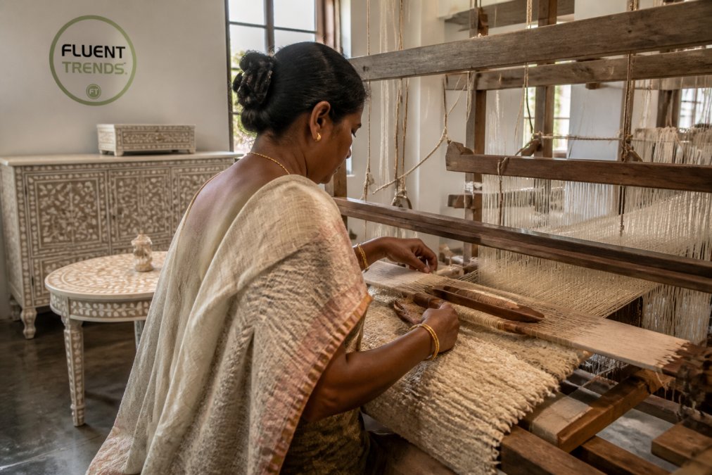 A woman artisan carefully weaving textured boucle fabric on a traditional wooden loom in a sunlit workshop, finished bone inlay furniture pieces visible in the background
