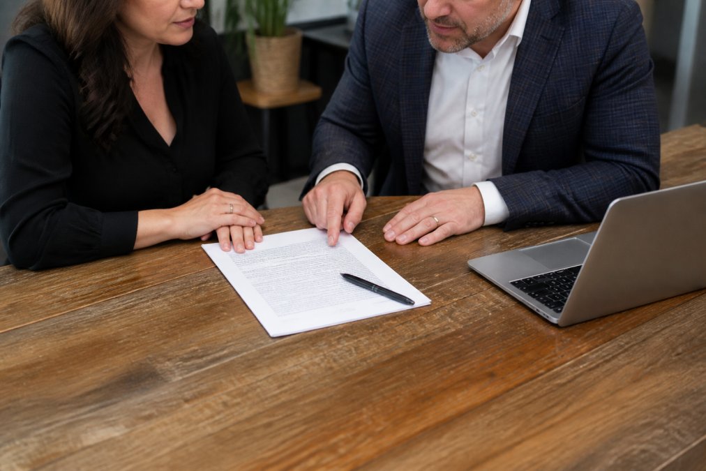 A business owner and an insurance broker reviewing a Broker of Record letter together at a wooden desk, with a laptop and a pen resting on the document