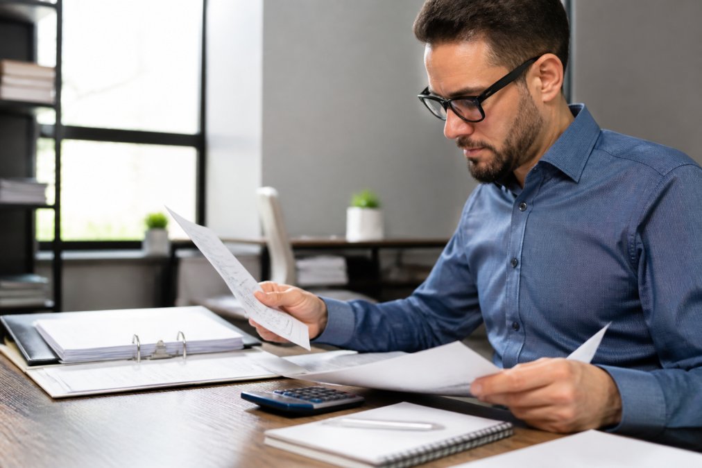 Business owner at desk reviewing payment processing statements and contract documents with a calculator and notepad visible, natural office lighting