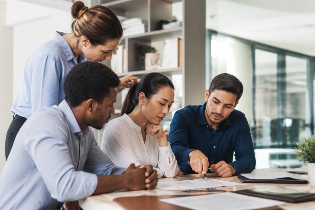 Small business team gathered around a computer monitor, collaborating on payment system setup with one person pointing at the screen, modern office setting with natural lighting