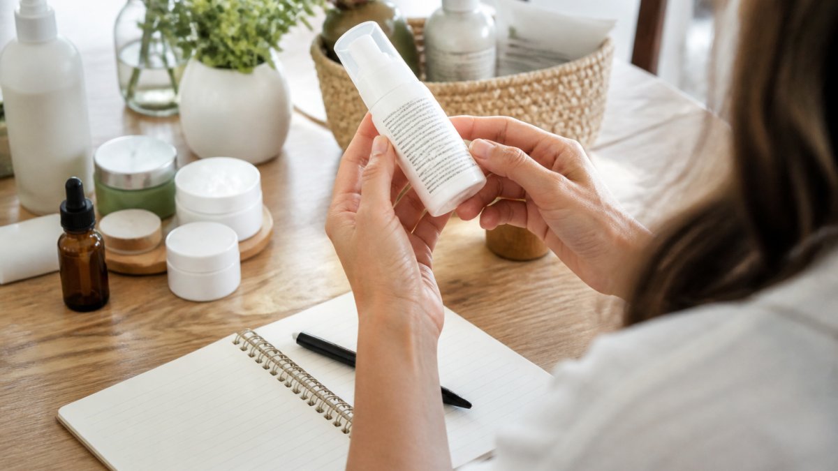 Hands examining a skincare bottle ingredient label to help with a switch to clean beauty while using a notebook