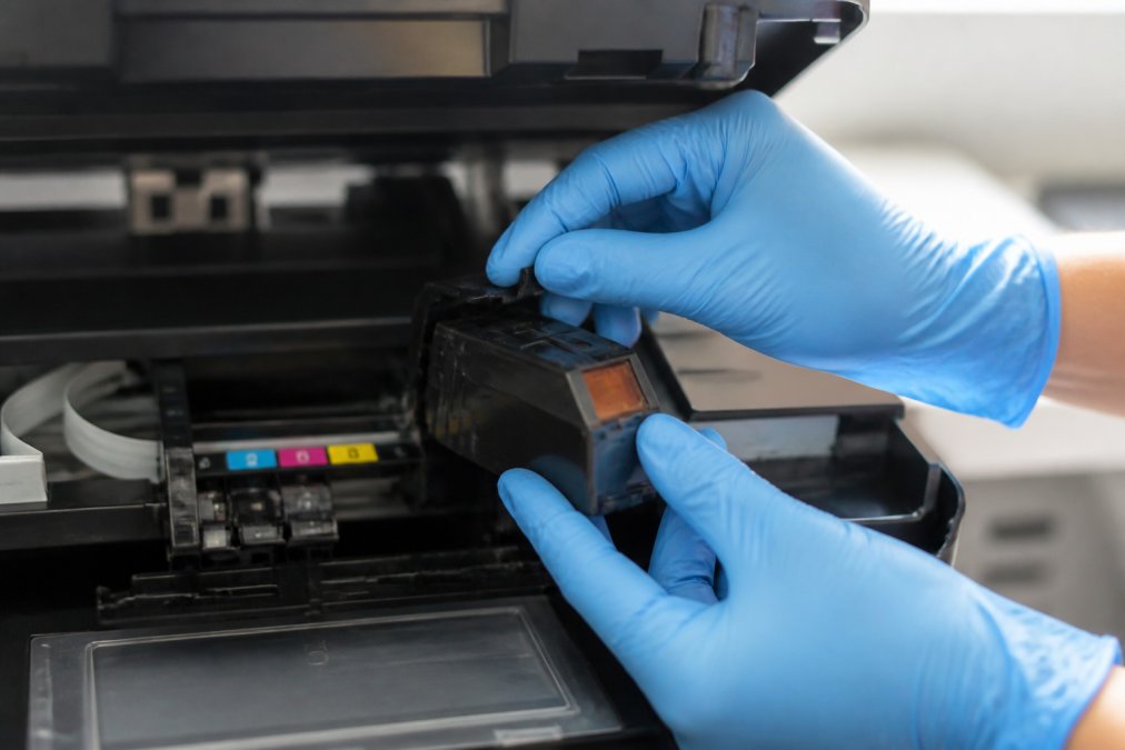 Close-up of hands wearing protective nitrile gloves carefully removing an OEM cartridge from a printer carriage, with the tab release mechanism visible and cartridge access area clearly shown in natural lighting