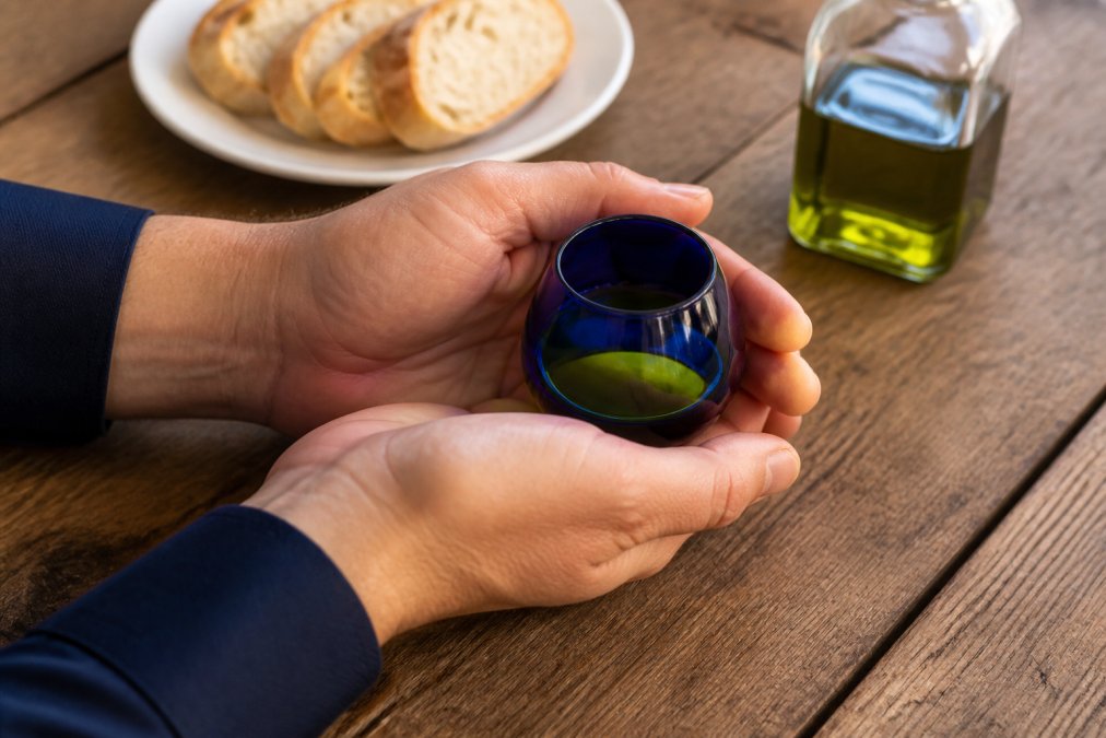 Close-up of hands cupping a small blue tasting glass of golden olive oil, warming it against the palm, with a white plate of sliced bread and a bottle of extra virgin olive oil on a rustic wooden table
