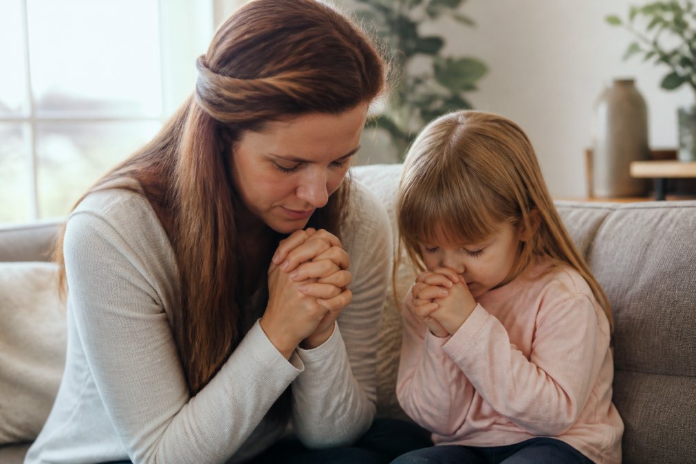A mother and young child sitting together on a couch with hands clasped, heads bowed in prayer, warm natural lighting from a nearby window, peaceful home setting