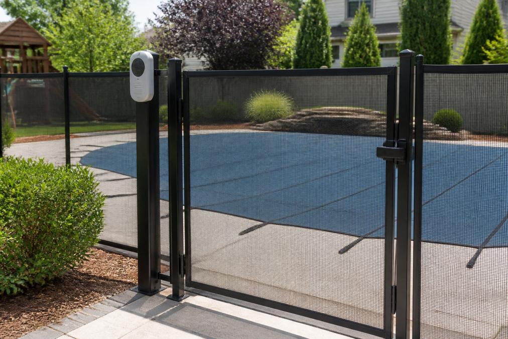 Residential backyard pool surrounded by secure four-sided safety fence with self-latching gate, pool alarm mounted on fence post, and safety cover visible on pool surface in afternoon sunlight