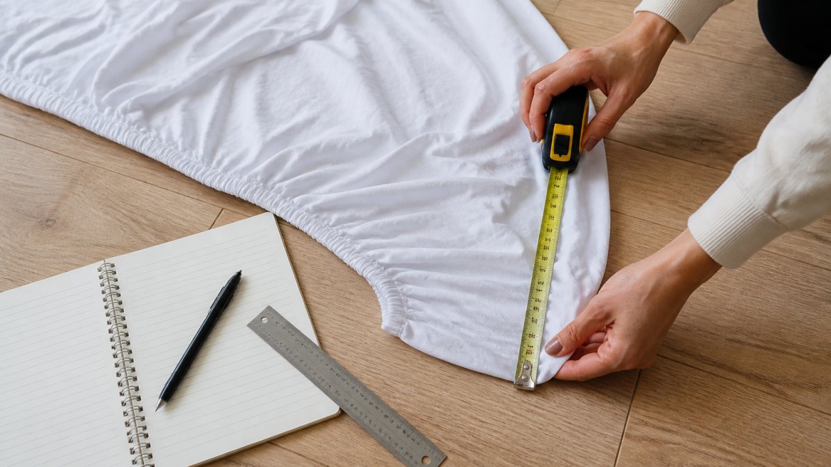 Close-up of hands using a retractable measuring tape to measure the corner of a white fitted sheet laid flat on a light wood floor, with the elastic edge visible and a notebook nearby