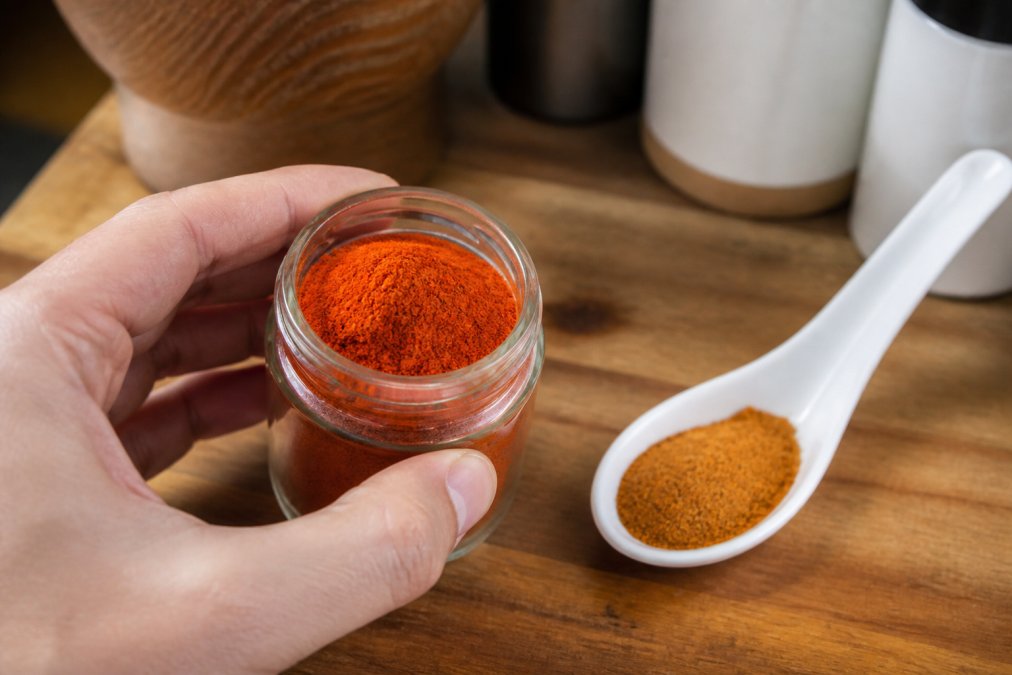 Close-up of a hand holding a small glass jar of vibrant red spice next to a white ceramic spoon of dull, faded spice powder on a wooden kitchen counter