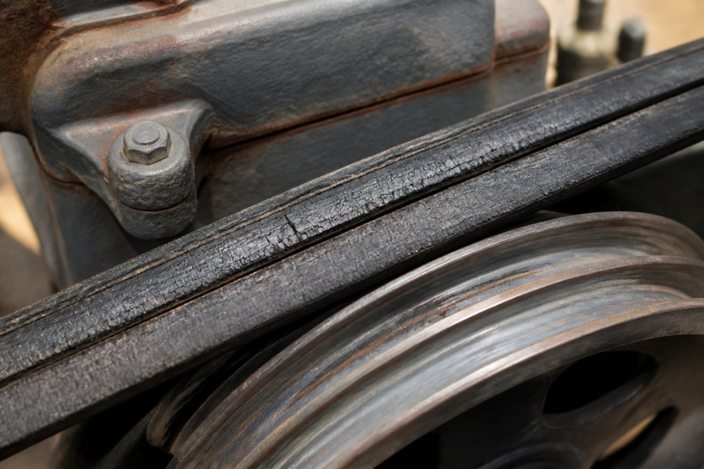 Close-up of a V-belt showing glazing, cracking, and surface degradation where it contacts a pulley system in industrial equipment
