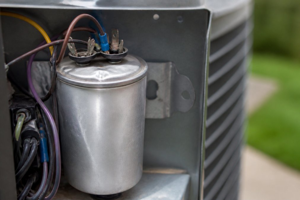 Close-up of a swollen, bulging AC capacitor mounted in an outdoor HVAC unit with visible deformation on the top casing and mounting bracket visible in background