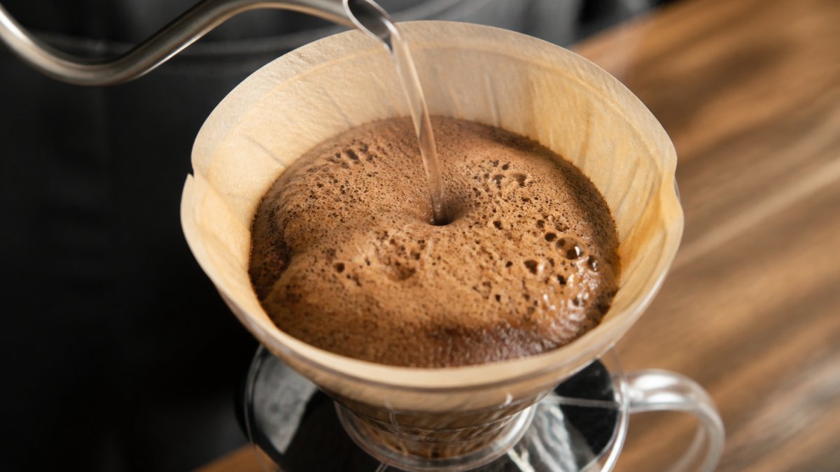 Close-up of a pour-over coffee brewer with hot water hitting fresh coffee grounds, showing a thick bubbly bloom rising in the filter