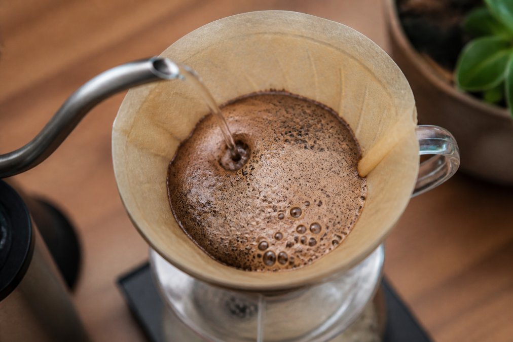 Close-up overhead shot of ground coffee in a glass pour-over brewer, showing a dramatic puffy bloom with bubbles and carbon dioxide escaping as hot water is poured from a gooseneck kettle