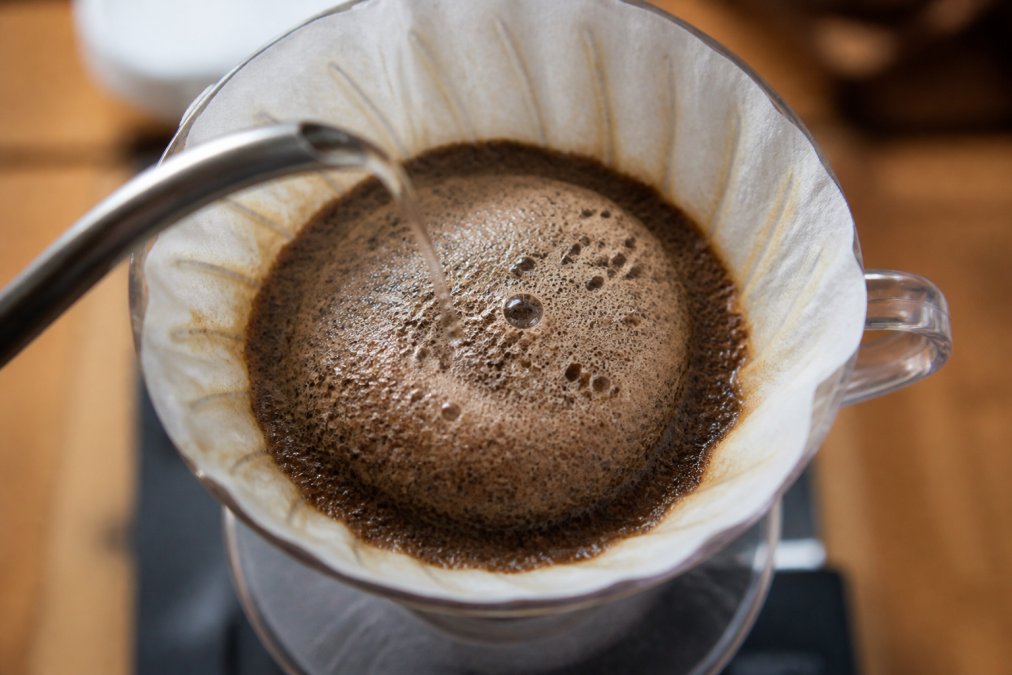 Close-up overhead shot of hot water being poured from a gooseneck kettle over coffee grounds in a glass pour-over dripper, a visible dome of CO2 bubbles rising upward from the dark, wet grounds under warm kitchen lighting