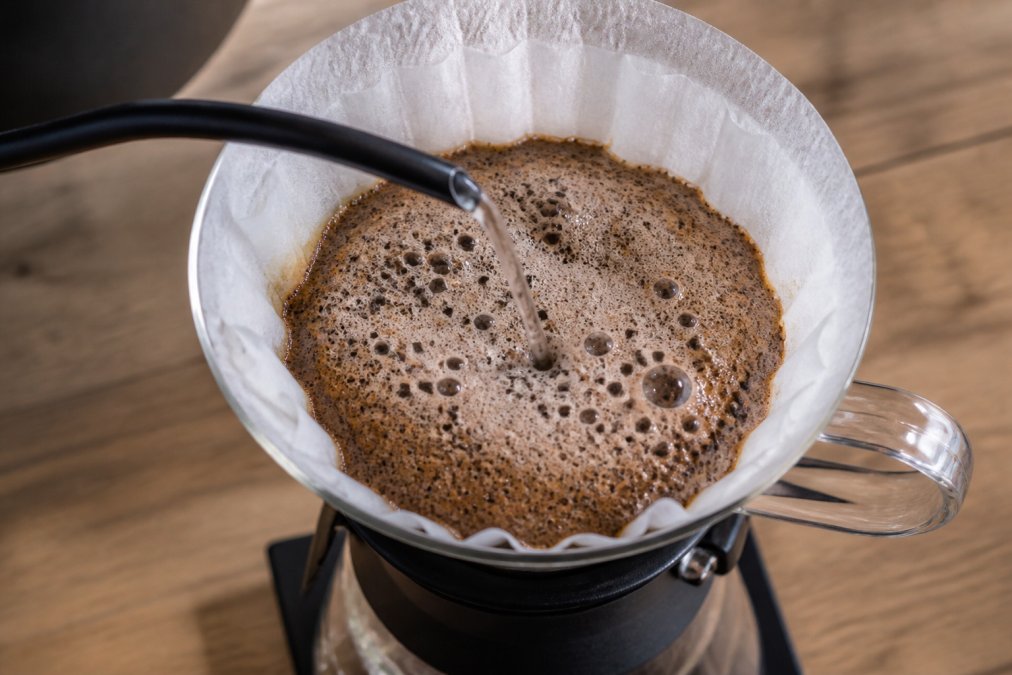 Close-up overhead shot of a pour-over coffee brewer with hot water hitting fresh ground coffee, showing a thick, bubbling bloom of foamy bubbles rising in the filter